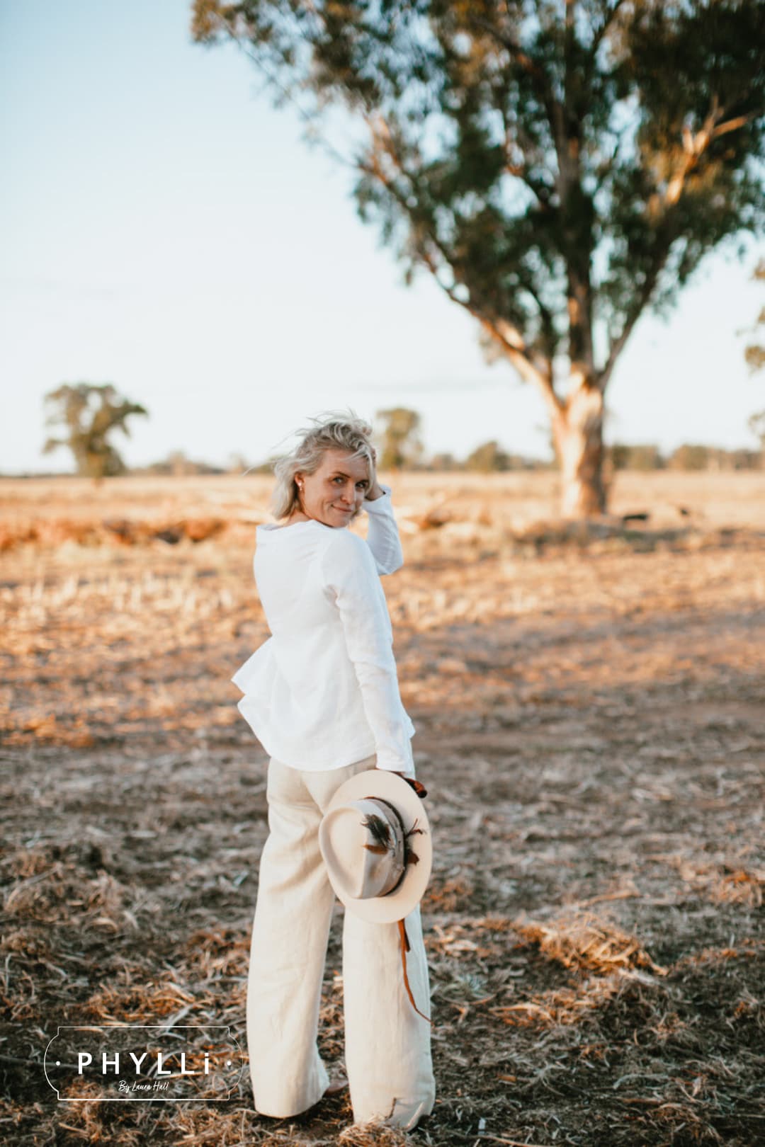 Woman holding the Roadie beige felt hat by PHYLLi Designs with feather cluster and suede ties in a dry rural paddock at sunset.