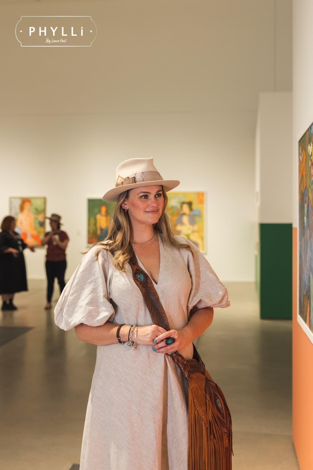 Laura Hall wearing the Roadie beige felt hat by PHYLLi Designs with distressed grosgrain band and feather trim in an art gallery.