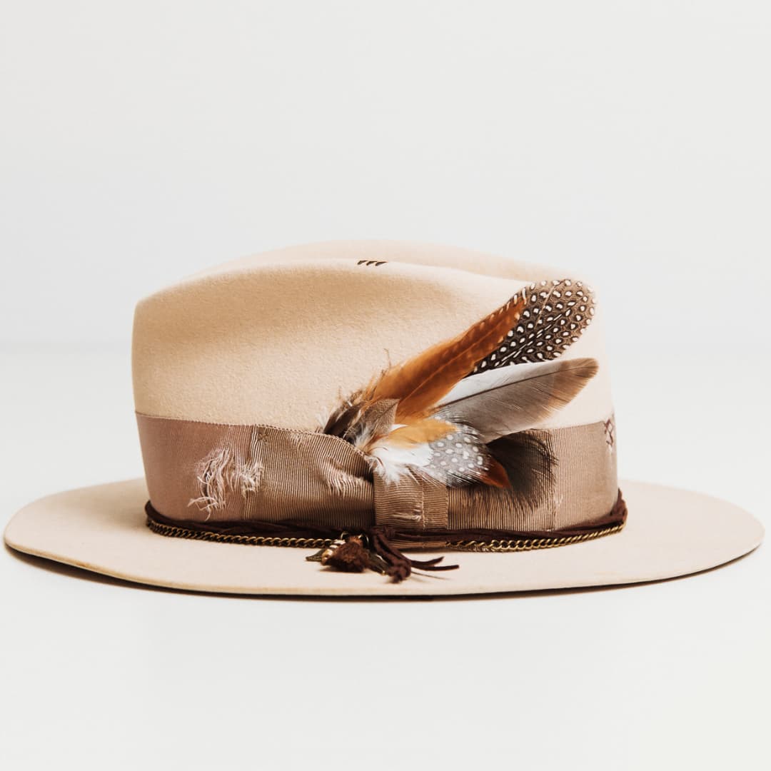 Left side view of the Roadie beige felt hat by PHYLLi Designs with distressed plaster grosgrain band, feather bunch and charm cluster.