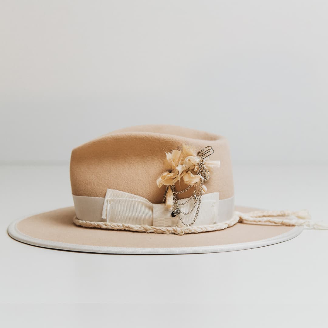 Left side view of the Rue De Seine beige felt hat by PHYLLi Designs with ivory grosgrain bow band, plaited sari silk brim trim and silver hat pin with tassel and charms.