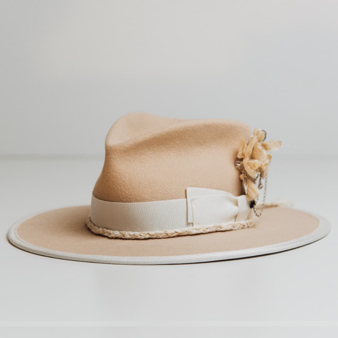 Front left angle of the Rue De Seine beige felt hat by PHYLLi Designs with ivory grosgrain bow band, plaited sari silk trim and silver chunky hat pin with charms.