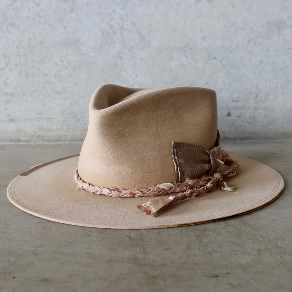 Front left angle of the sand distressed felt hat Ryoan Limited Edition by PHYLLi Designs with taupe Japanese grosgrain floating double bow and plaited sari silk trimmings in tonal pinks.