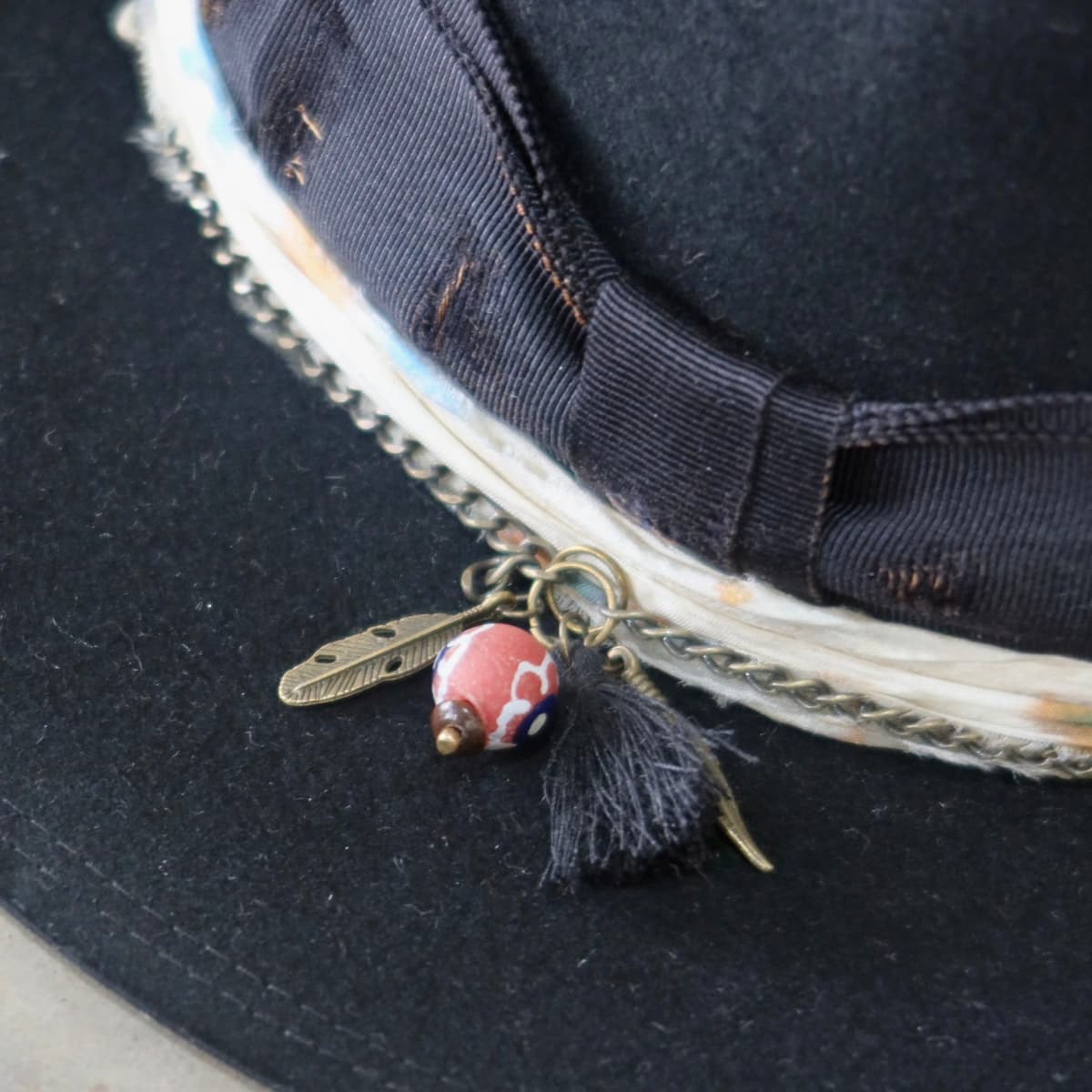 Close up detail of the Saint felt hat showing charm cluster with black tassel, bronze feather charm and recycled glass Ghana bead pendant.
