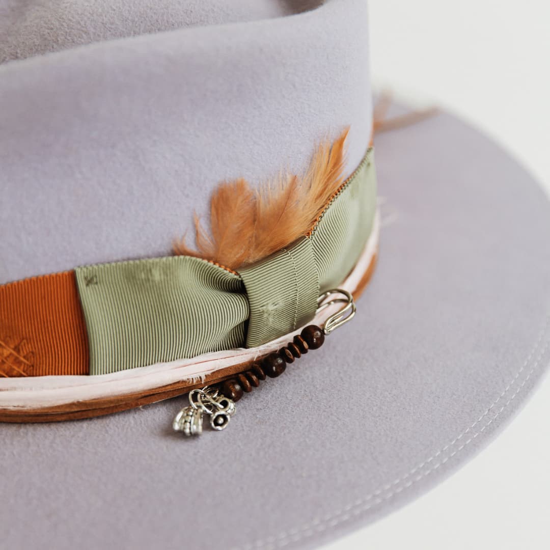 Close up detail of Santa Monica Lilac felt hat by PHYLLi Designs showing oil green bow, rust grosgrain band, tan chicken feathers and silver hat pin.