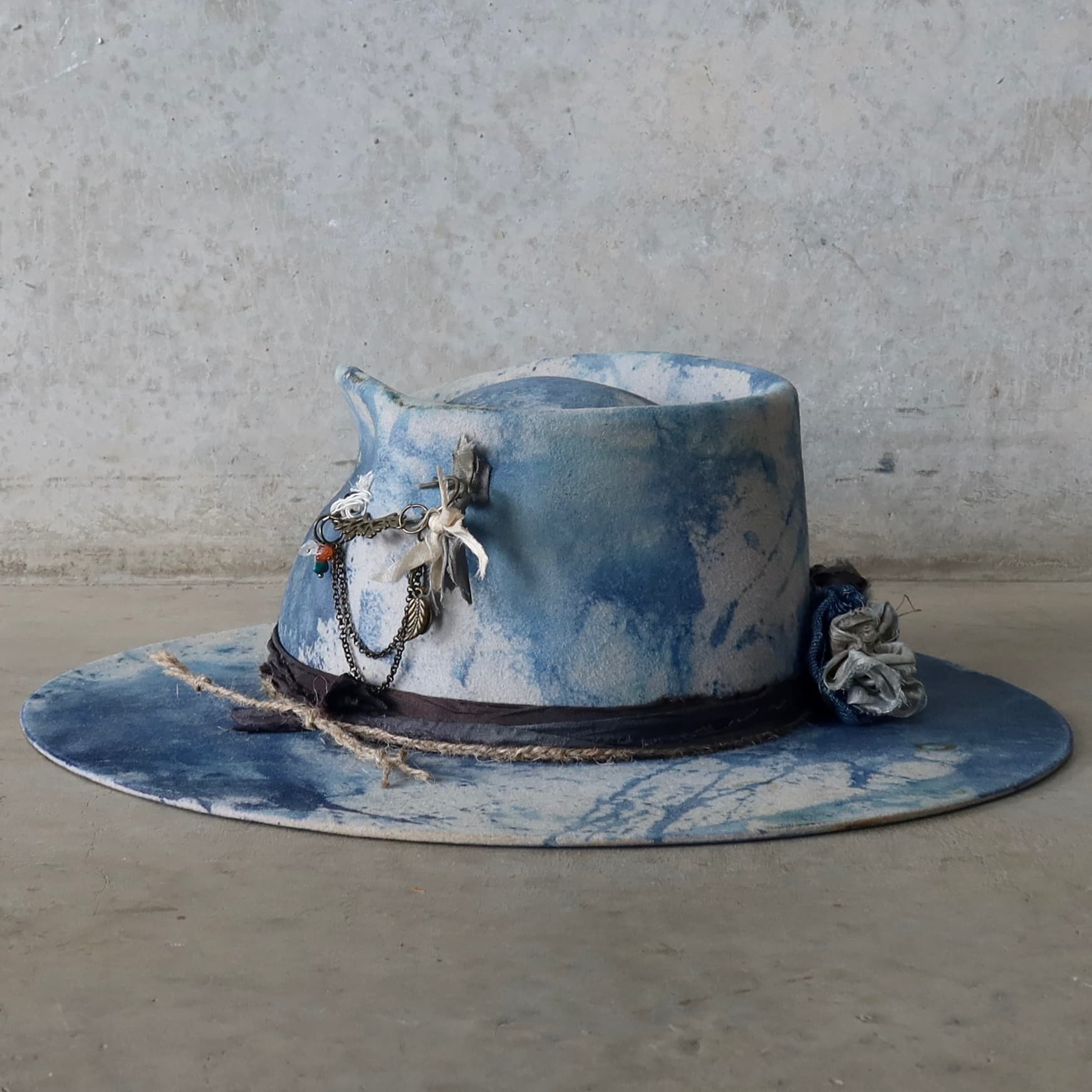 Back left angle of the dove grey indigo tie dye felt hat Shibori Limited Edition by PHYLLi Designs with bronze hat pin and charm chains.