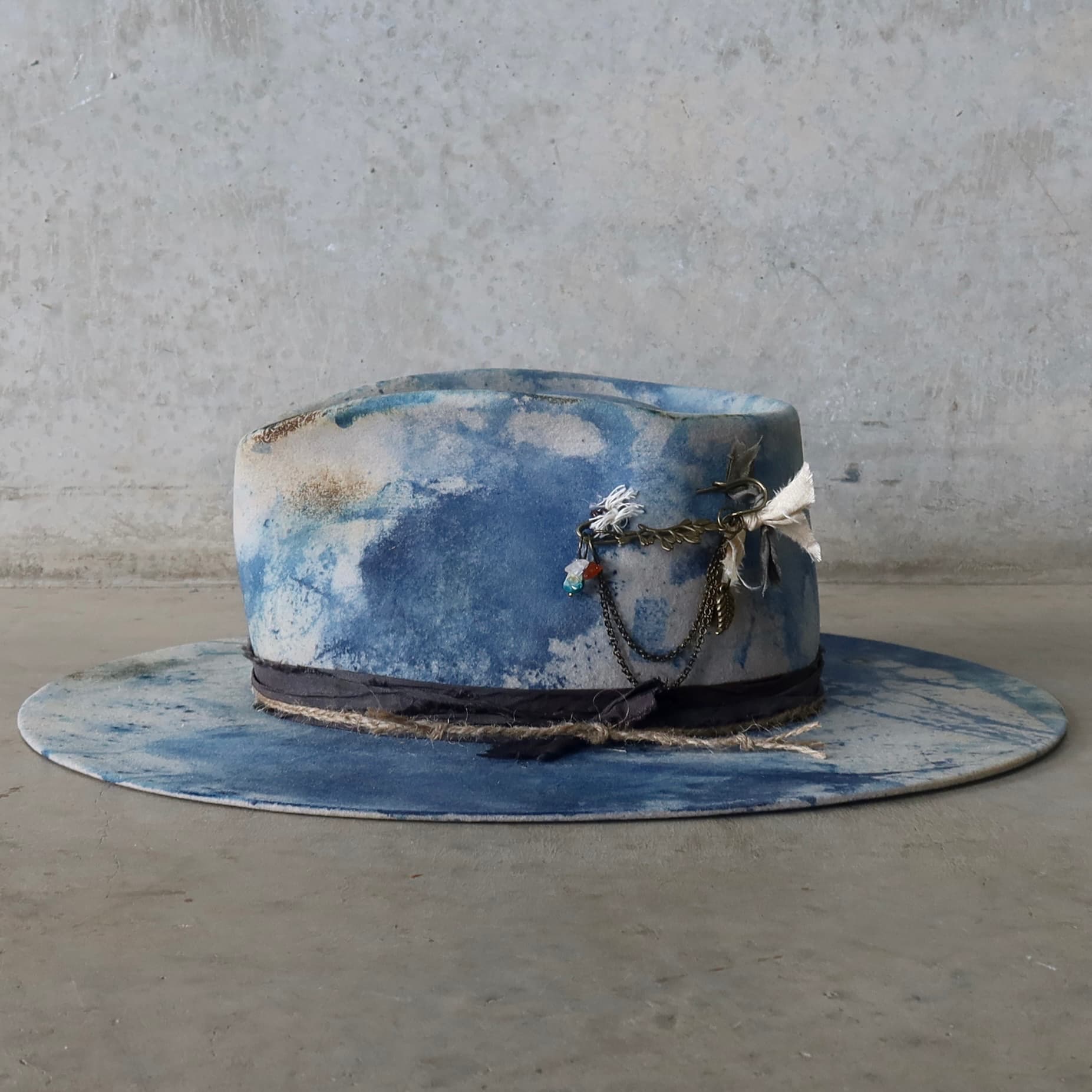 Left side view of the dove grey indigo tie dye felt hat Shibori Limited Edition by PHYLLi Designs with bronze hat pin featuring chains, charms and quartz crystals.