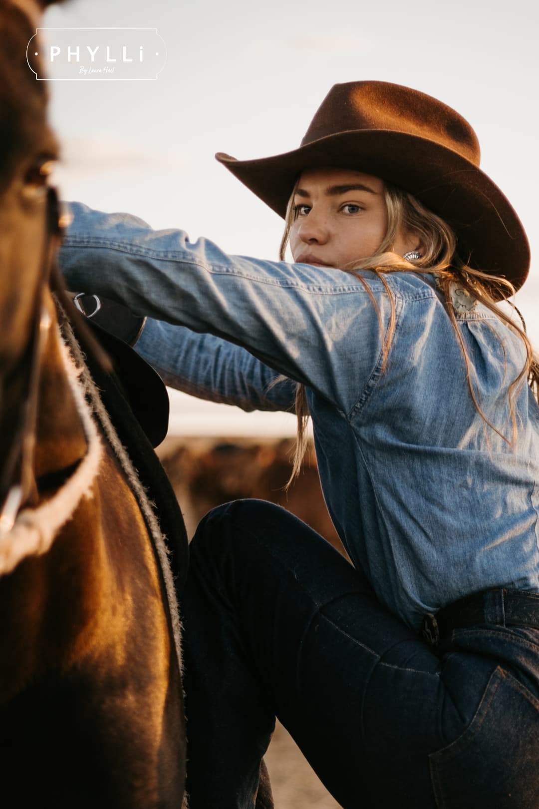 Model mounting a horse wearing the chocolate cowboy hat Spicer by PHYLLi Designs with chocolate grosgrain band and leather tie back in an outback paddock.