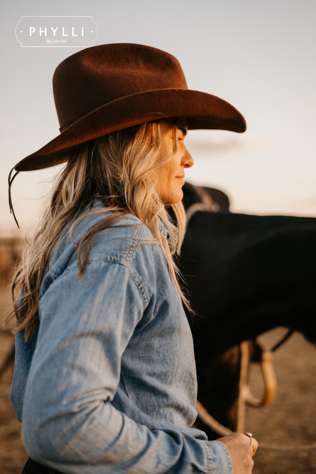 Woman wearing the chocolate cowboy hat Spicer by PHYLLi Designs with leather tie back band beside a saddled horse at sunset.