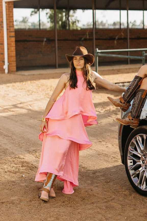 Woman wearing the PHYLLi Spicer chocolate cowboy hat with a pink tiered dress, standing beside a car on a dirt road.