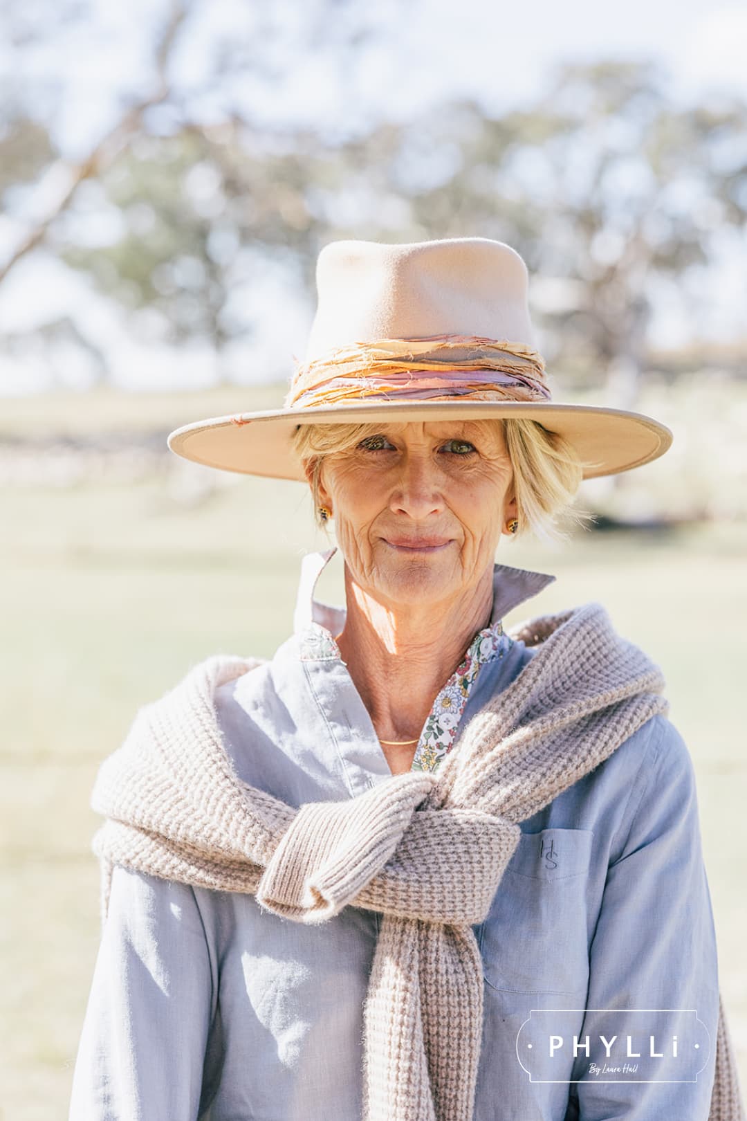 Older woman wearing the Sunset sand felt hat by PHYLLi Designs with layered sunset-toned sari silk trim and a soft knit draped over her shoulders.