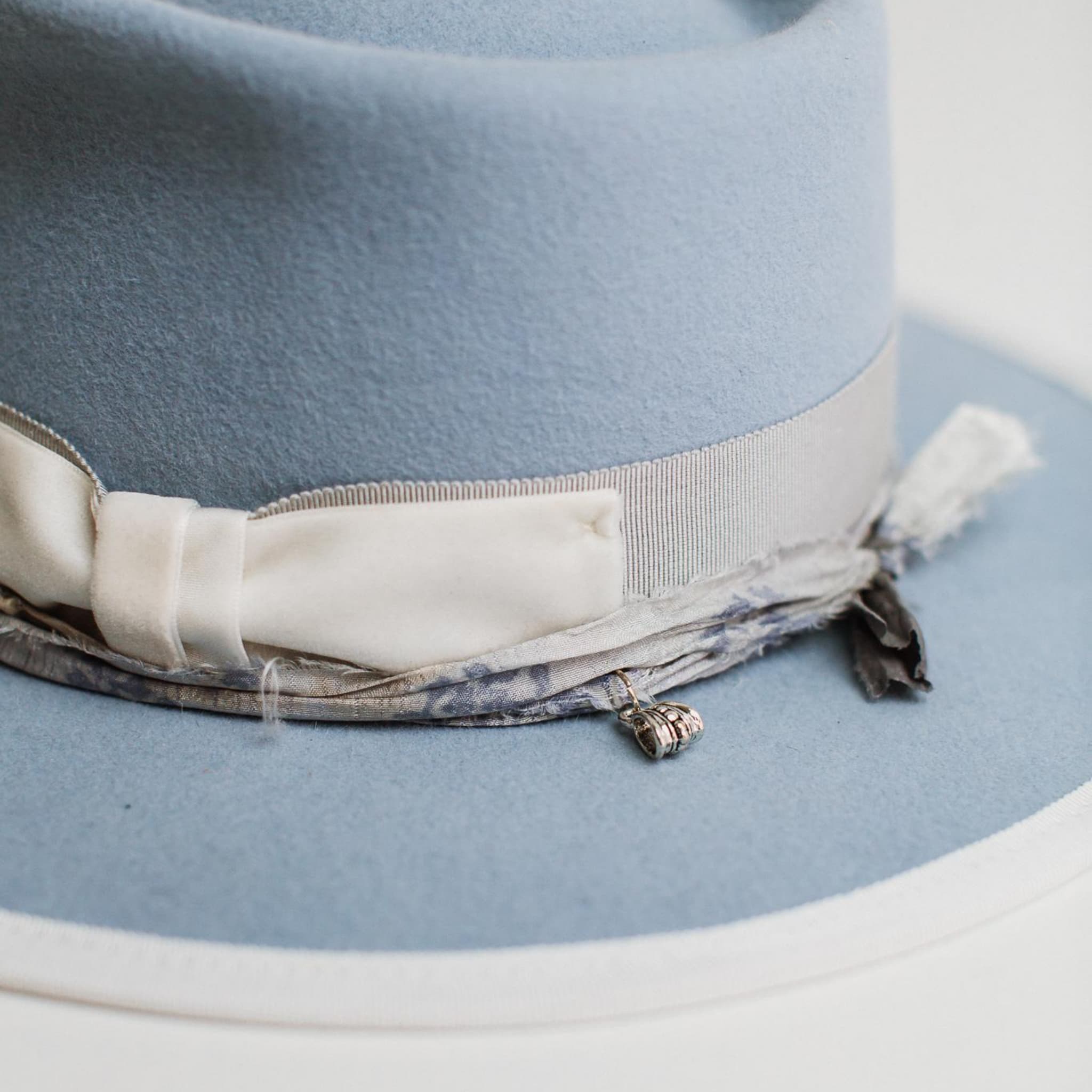 Close up detail of The Arrow crystal blue felt hat by PHYLLi Designs featuring dove grosgrain band, ivory velvet bow, dusty blue sari silk trim and silver pendant charm.