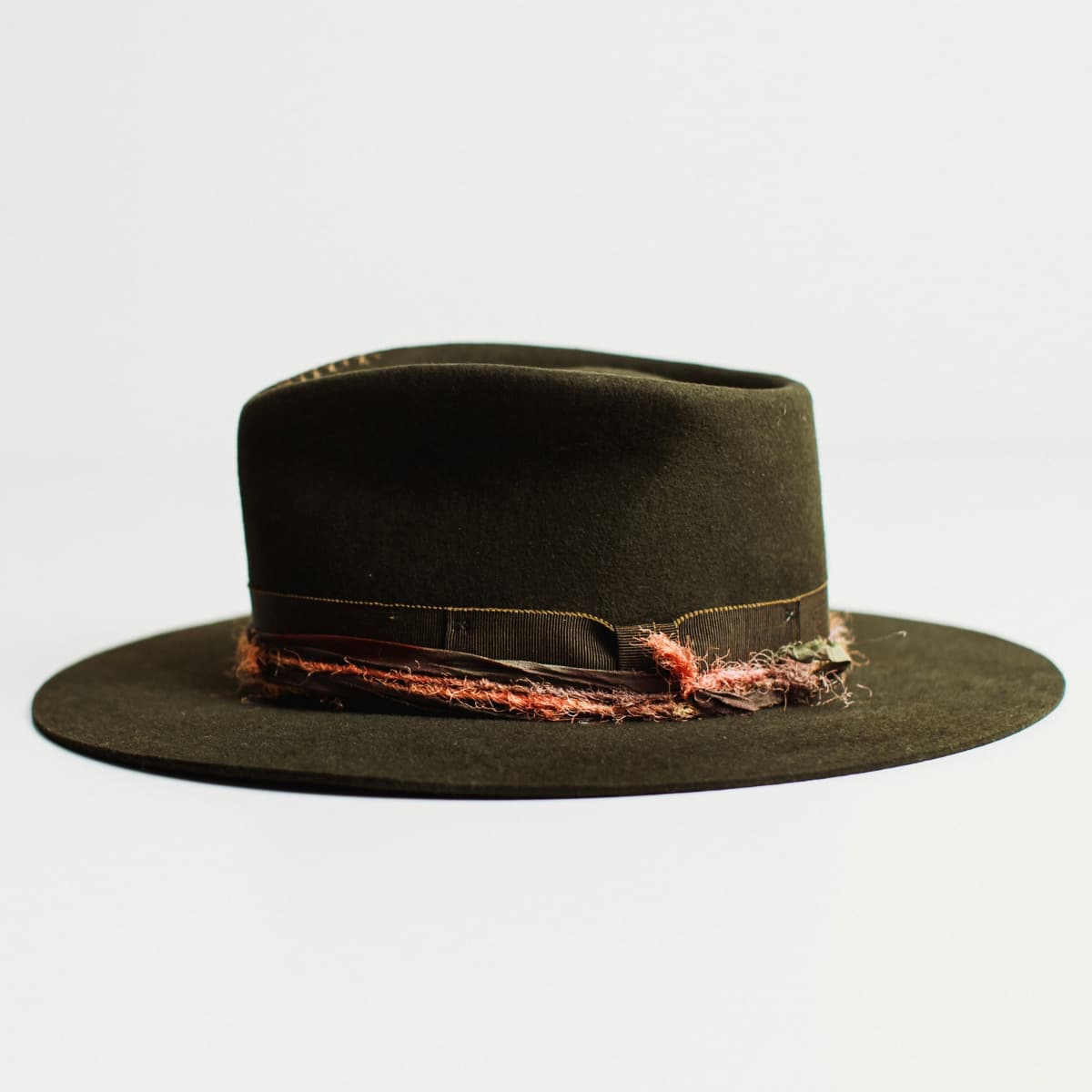 Left side view of dark olive felt hat with distressed layered band on a light grey background.
