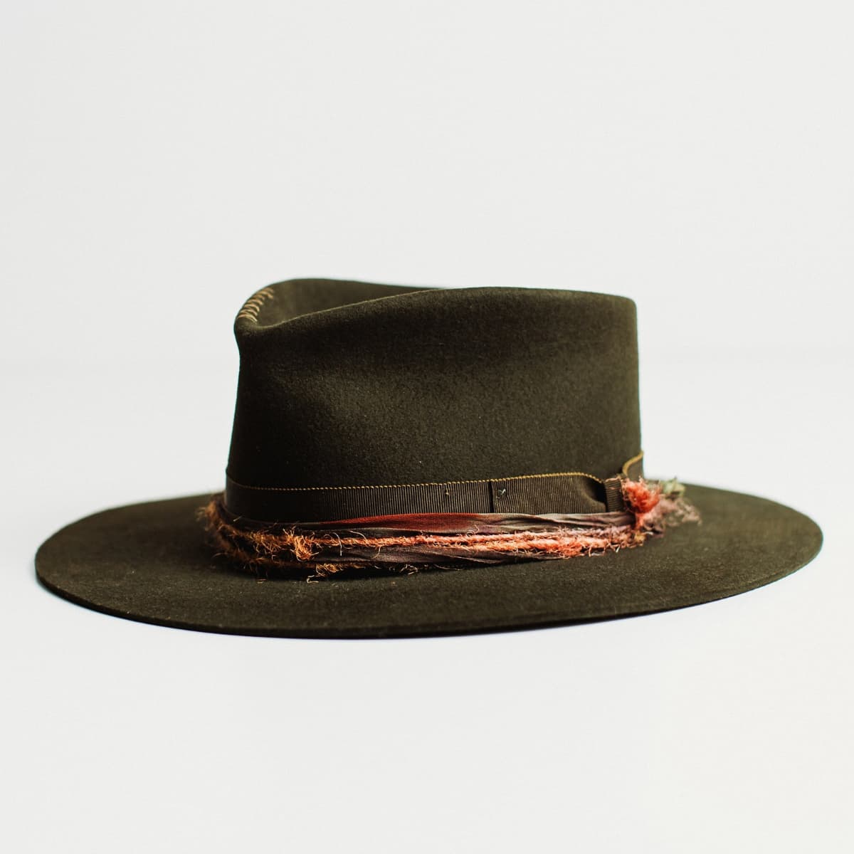 Front left angled view of dark olive felt hat with distressed layered band by PHYLLi Designs on a light grey background.