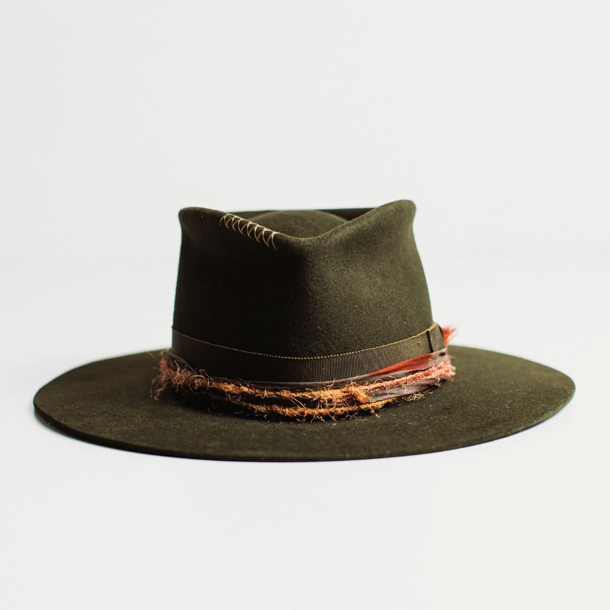 Front view of dark olive felt hat with stitched crown detail and distressed layered band by PHYLLi Designs on a light grey background.