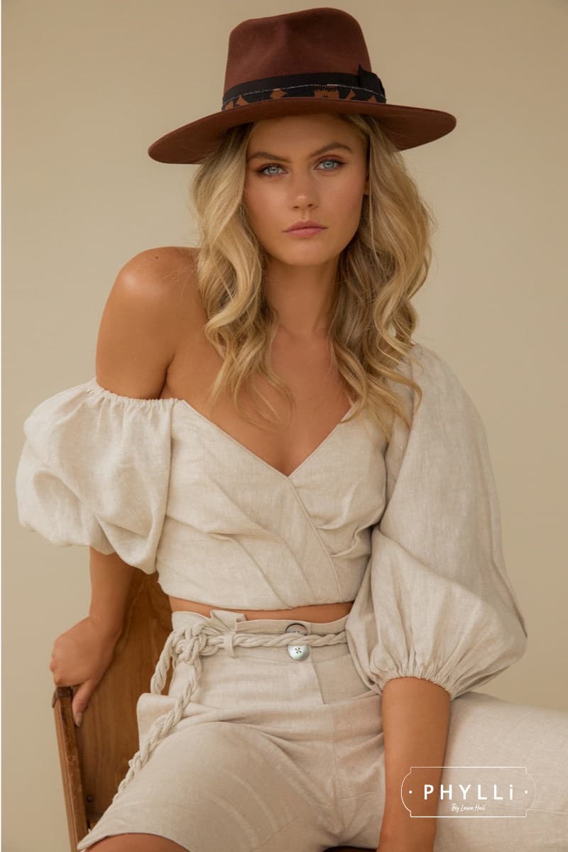Woman wearing a russet felt hat styled with a beige outfit while seated on a wooden chair.