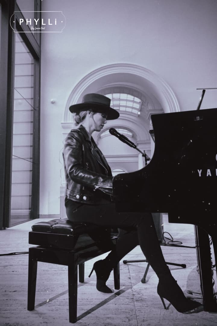 Delta Goodrem playing piano while wearing a russet felt hat by PHYLLi Designs in a room with large windows.
