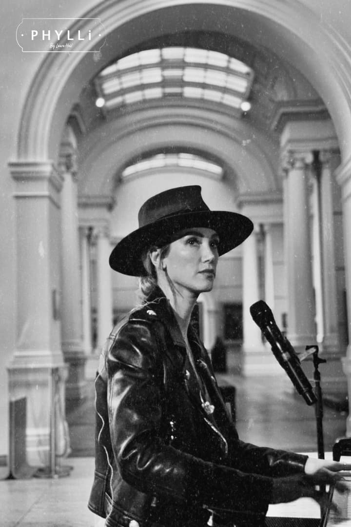 Delta Goodrem wearing a russet felt hat by PHYLLi Designs while seated at a piano in a grand hall.