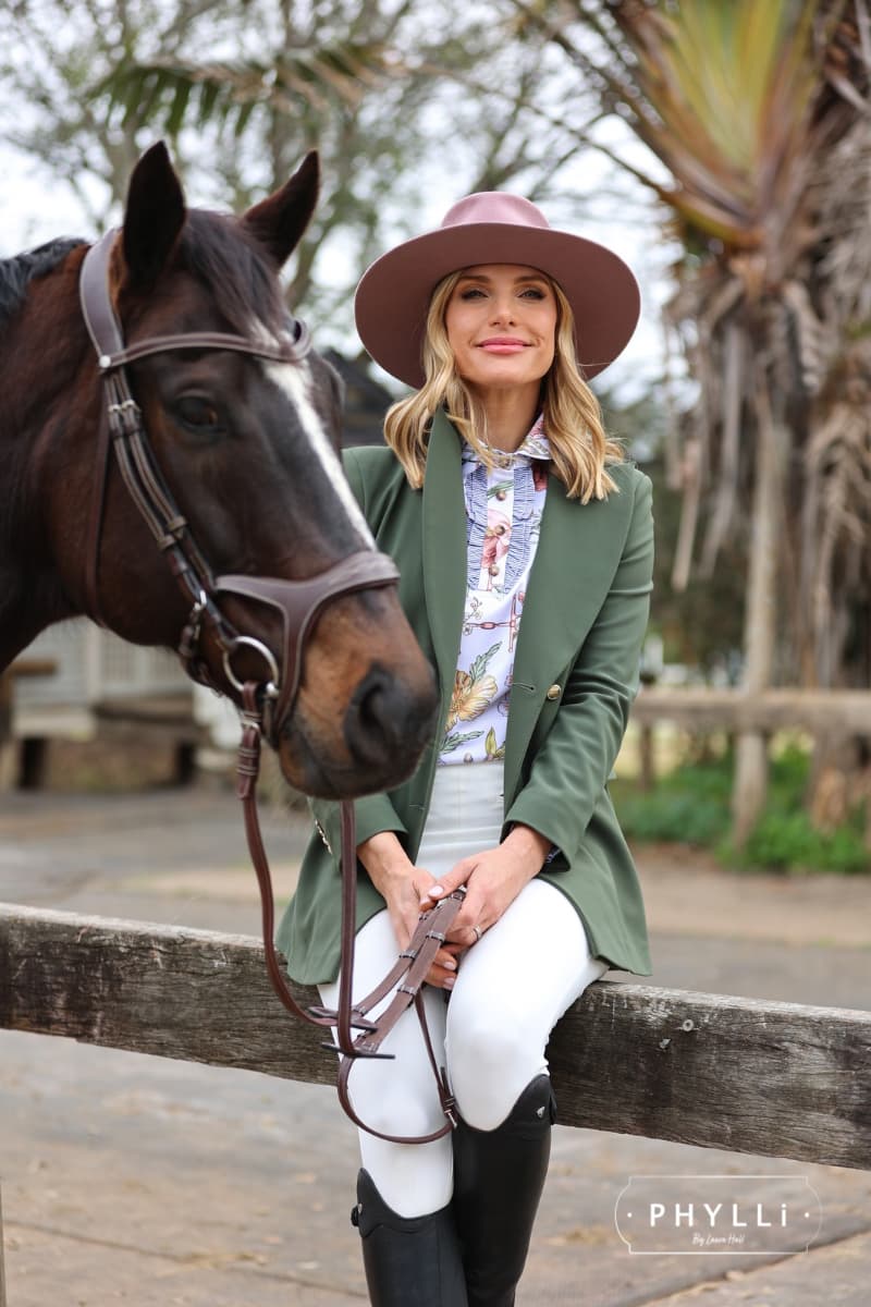 Woman in equestrian outfit wearing The Blush pink felt hat sitting on fence next to horse.