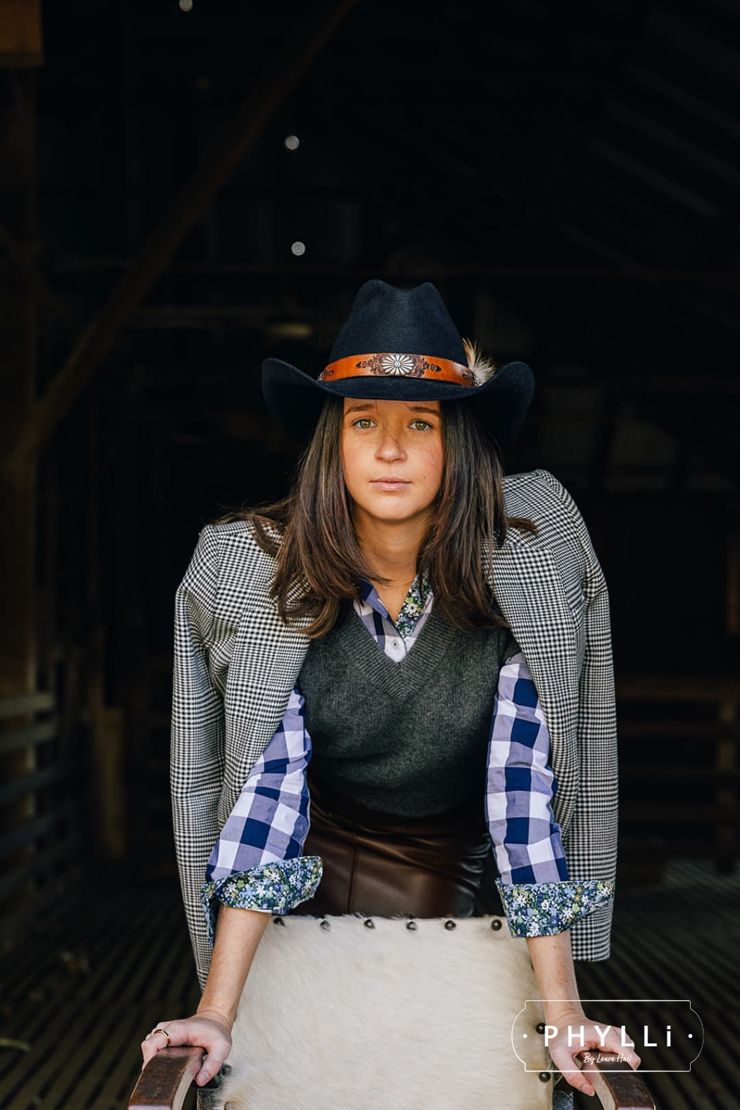 Model wearing The PHYLLi dark navy felt hat by PHYLLi Designs with Buffalo Girl Rugged Desert tie back leather band and feather trim inside a rustic timber barn interior.