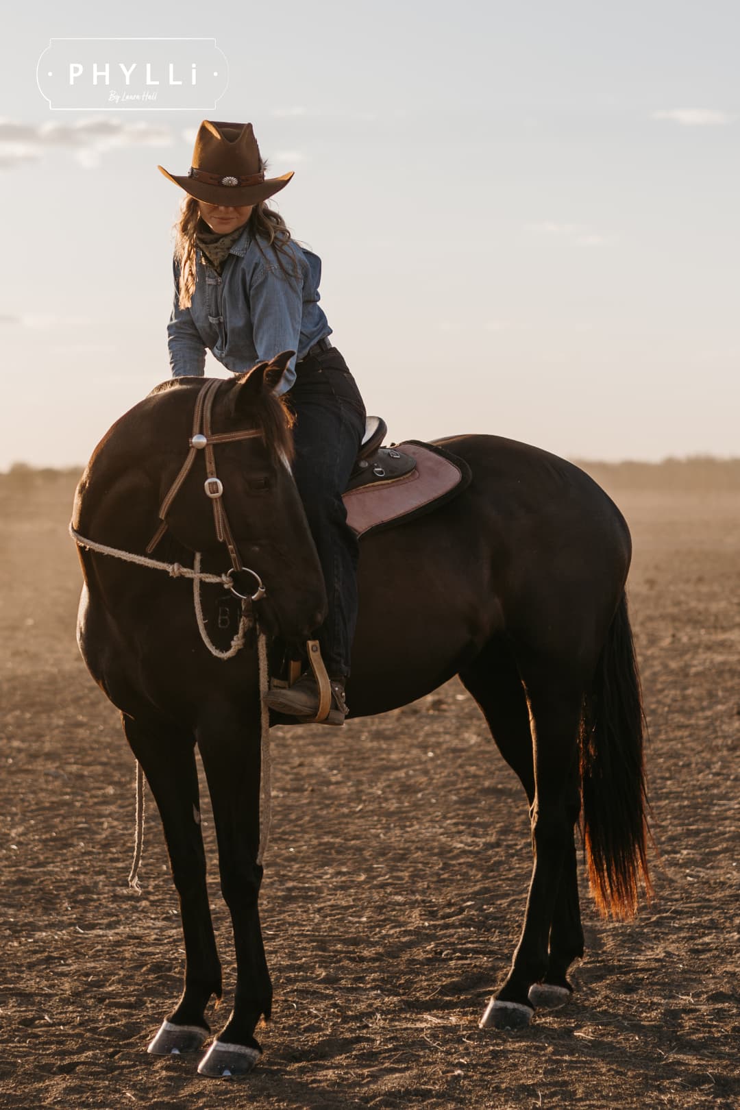 Cowgirl wearing the Caramel cowboy hat The PHYLLi Caramel by PHYLLi Designs with Buffalo Girl Western floral tie back hat band riding horseback in an open country landscape.