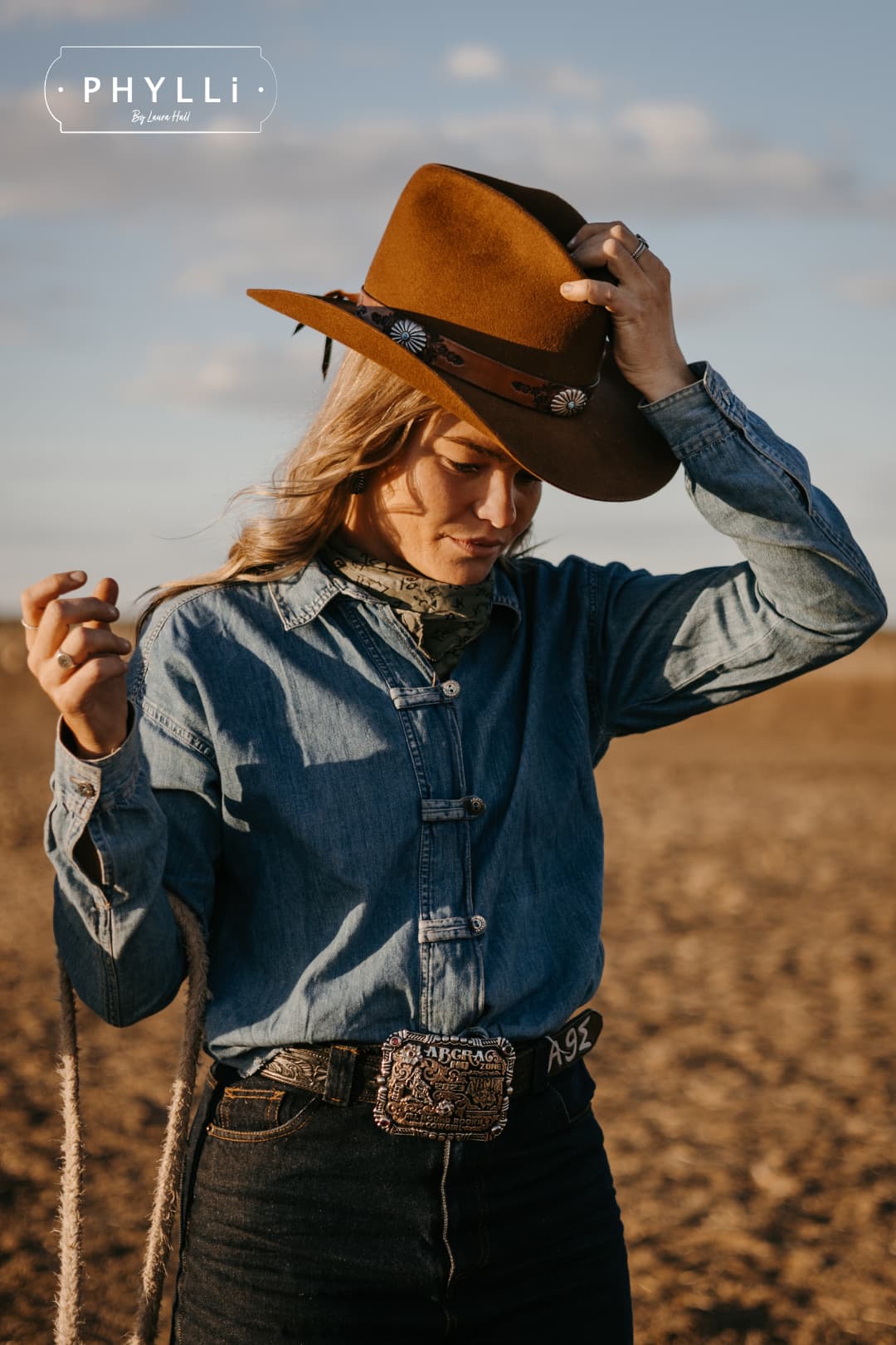 Woman wearing the Caramel cowboy hat The PHYLLi Caramel by PHYLLi Designs with Buffalo Girl Western floral tie back hat band in an open rural paddock at golden hour while adjusting the brim.