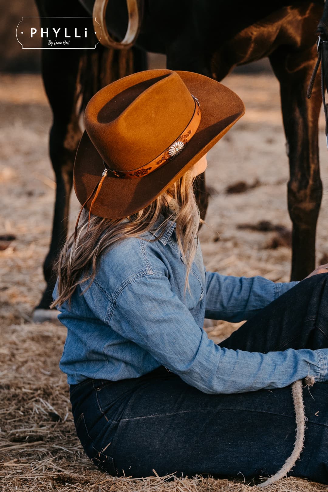 Cowgirl wearing the Caramel cowboy hat The PHYLLi Caramel by PHYLLi Designs with Buffalo Girl Western floral tie back hat band sitting in a rustic paddock beside a horse.
