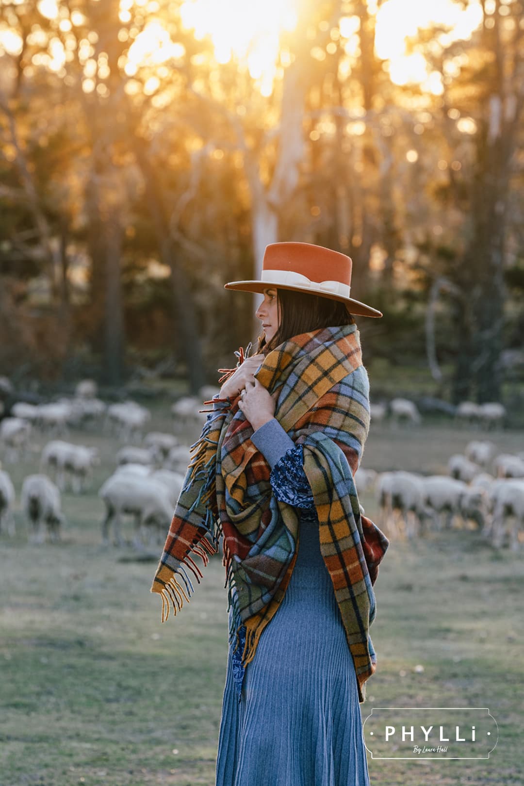 Woman wearing the Producer hat in spiced pumpkin with a plaid shawl standing in a field with sheep.