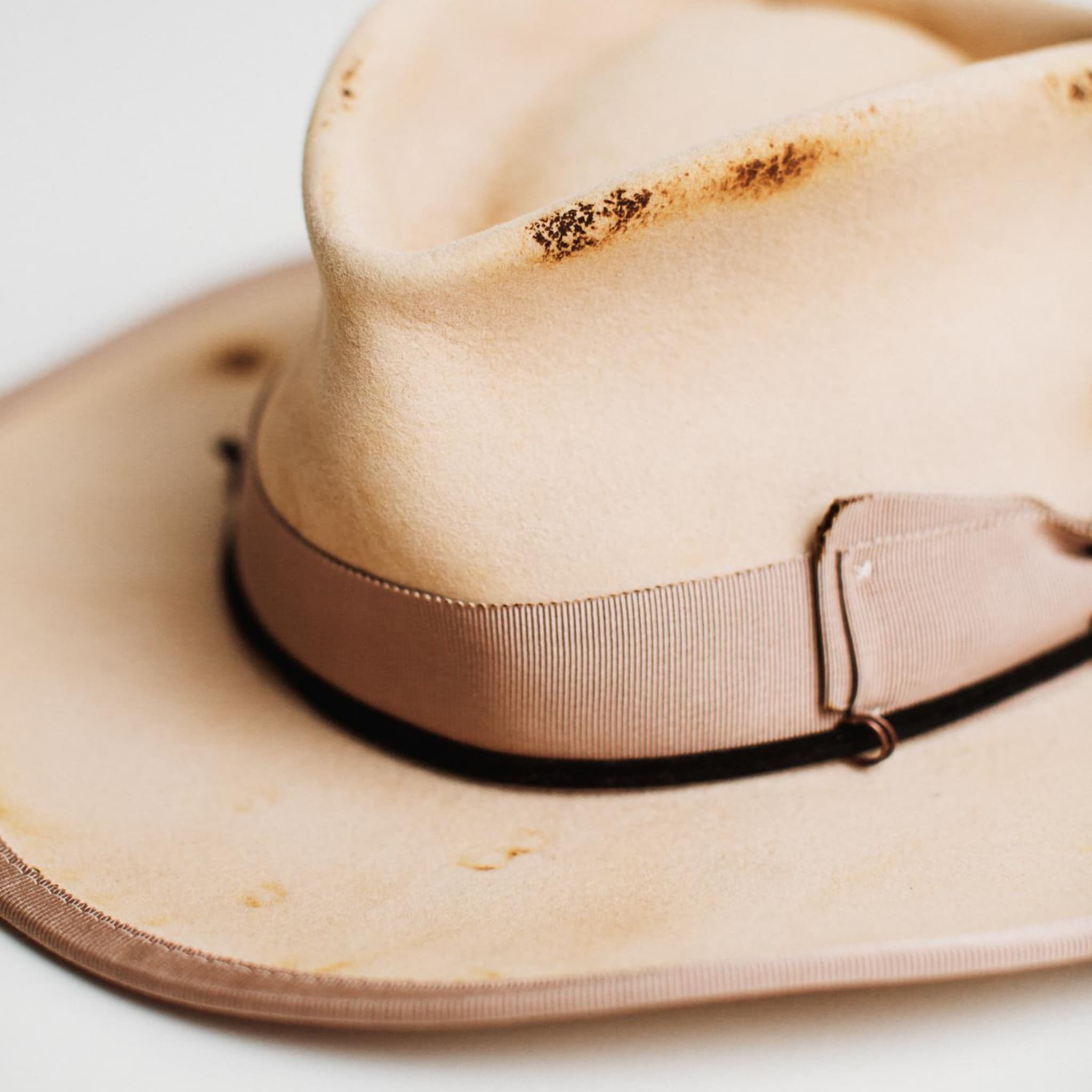 Close-up detail of Ringleader beige distressed felt hat by PHYLLi Designs showing plaster grosgrain double bow band, distressed felt finish and black suede string with bronze O rings.