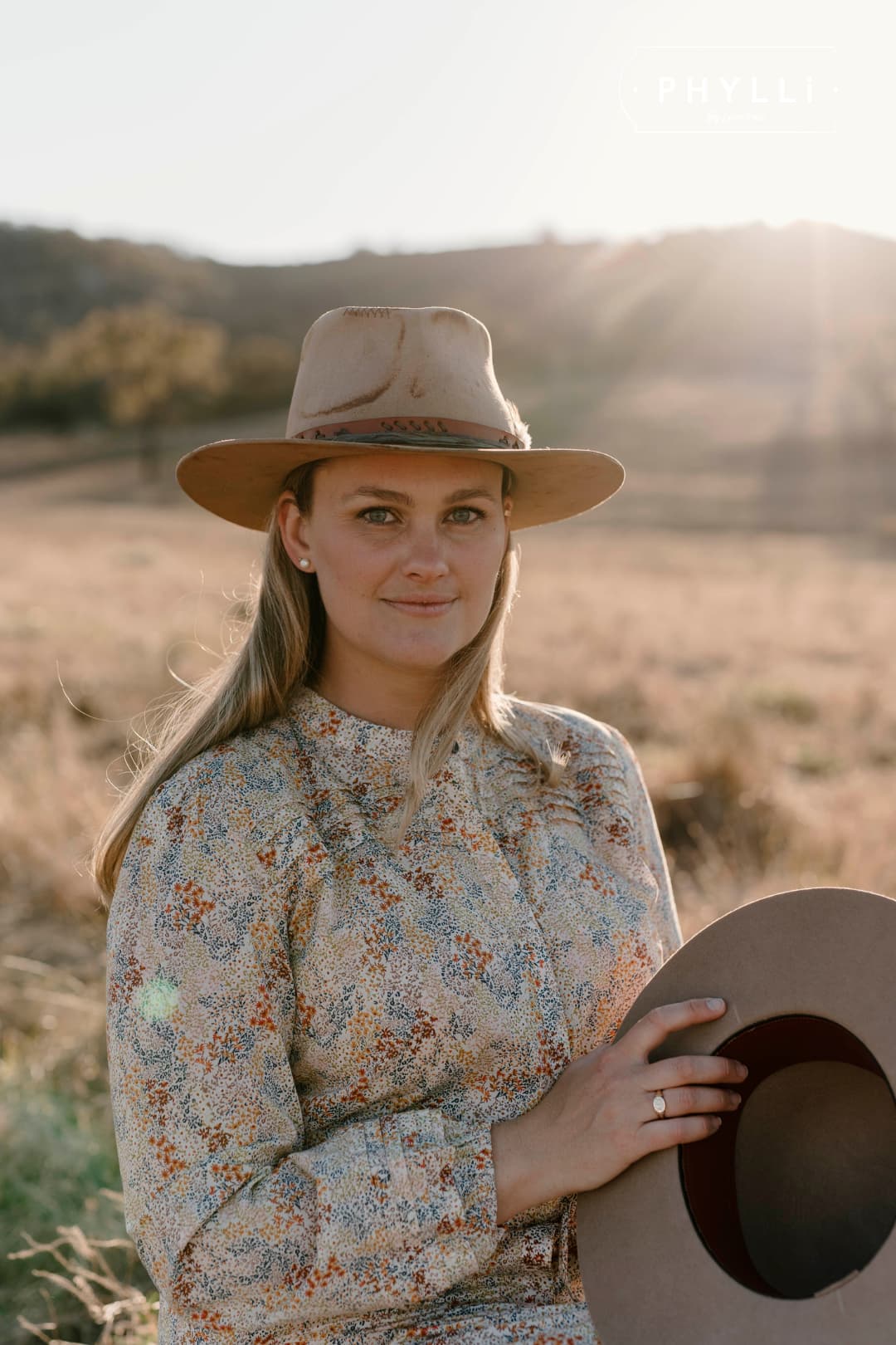Laura Hall, founder of PHYLLi Designs, wearing The Safari distressed Sahara felt hat and holding another hat in rural landscape.