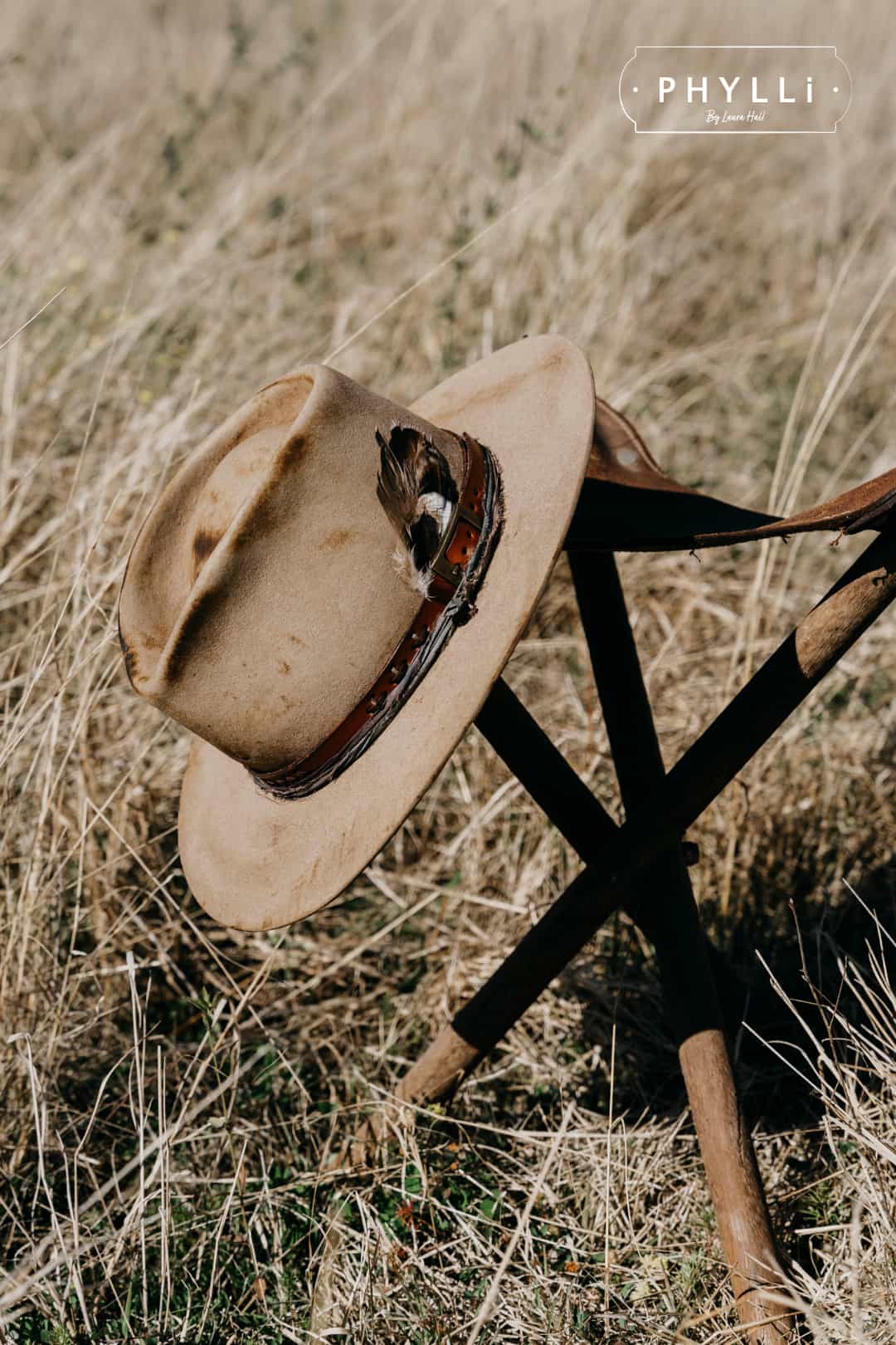 The Safari distressed Sahara felt hat by PHYLLi Designs resting on a rustic wooden stool in dry grass.