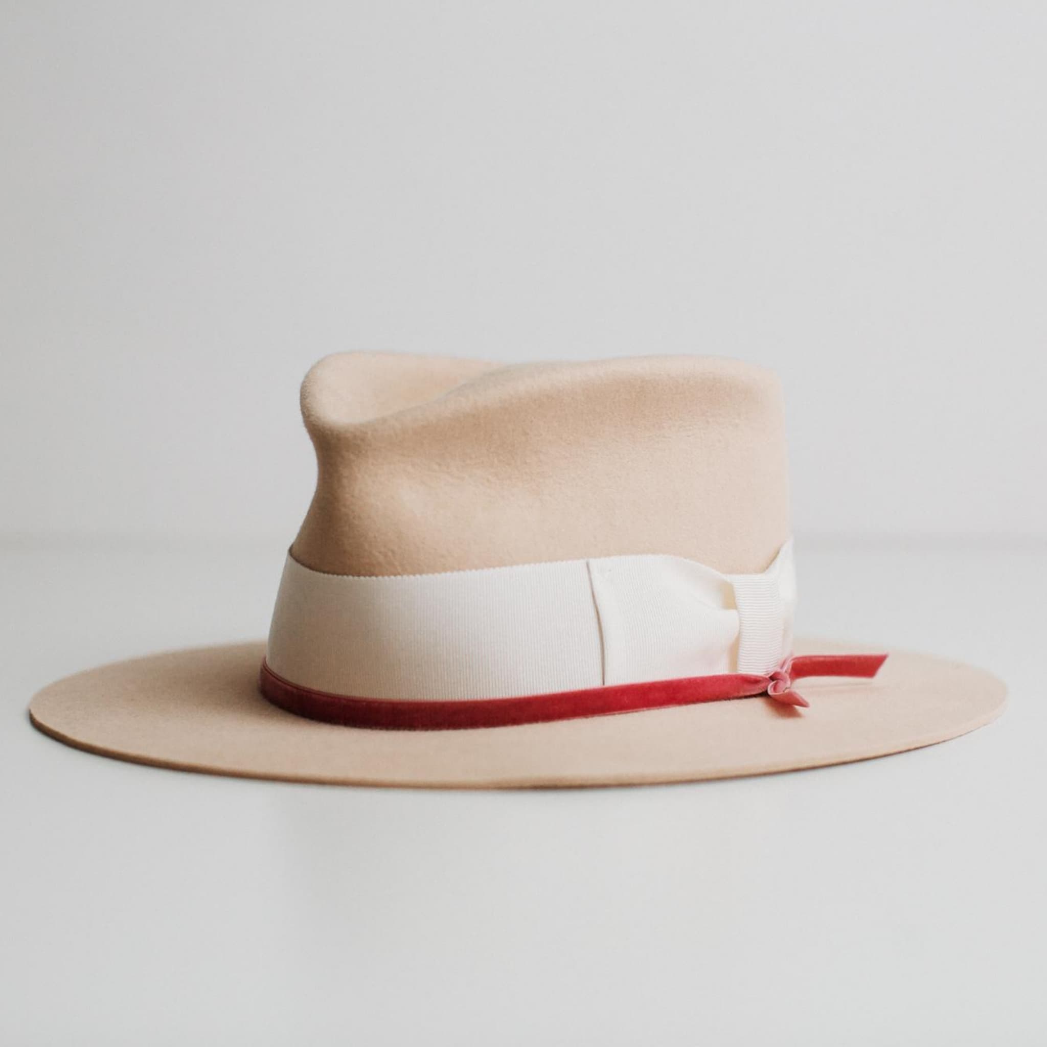 Angled view of beige felt hat with ivory band and pink velvet ribbon by PHYLLi Designs on a light grey background.