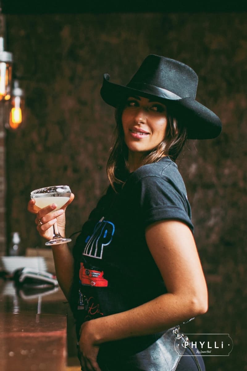 Woman wearing a black felt cowboy hat by PHYLLi Designs holding a cocktail in a dimly lit setting.