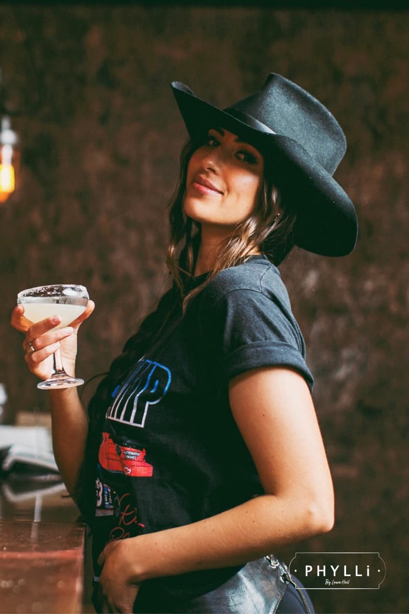 Woman wearing a black felt cowboy hat by PHYLLi Designs holding a drink with a blurred background.