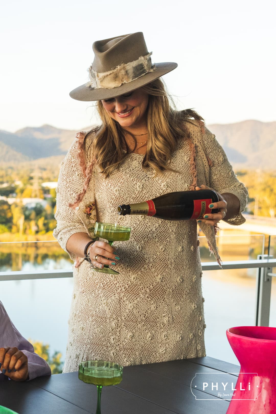 Laura Hall, founder of PHYLLi Designs, pouring champagne while wearing the Wabi Limited Edition Sahara felt hat with Wabi Sabi patch band on a balcony overlooking mountains.