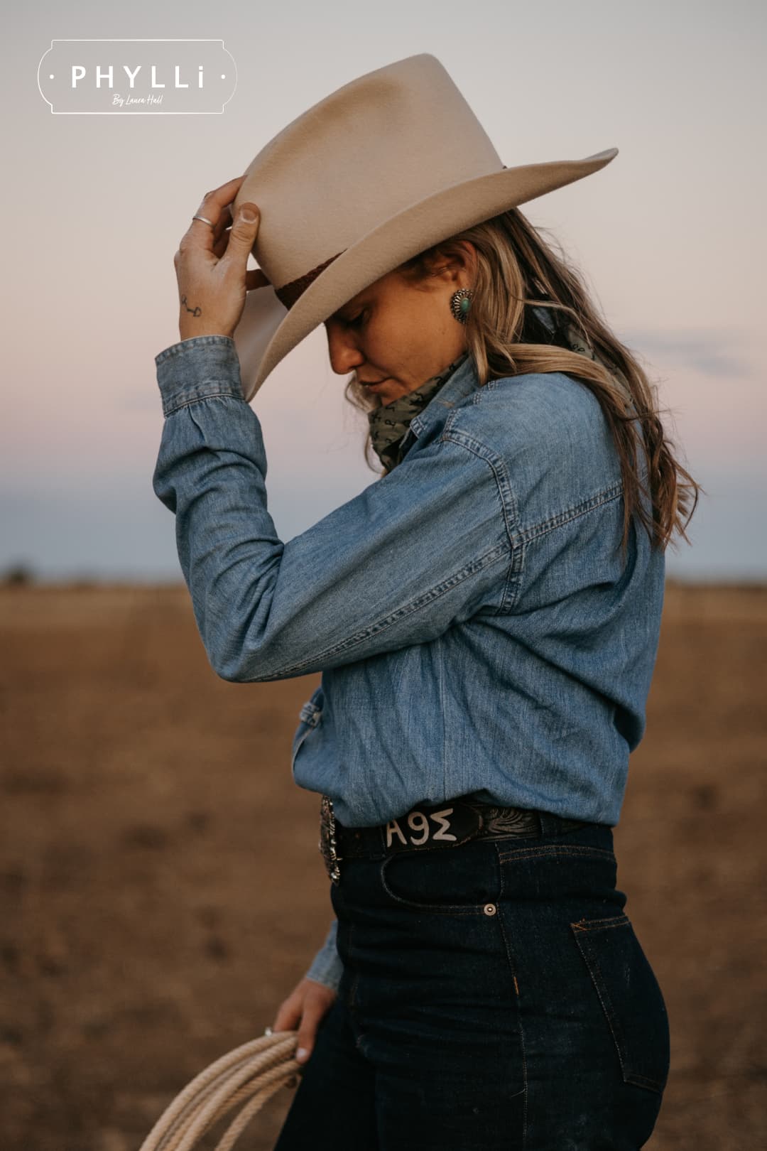 Model wearing the beige cowboy hat Wheeler Beige by PHYLLi Designs with chocolate leather hat band while adjusting the brim in a rural paddock setting.