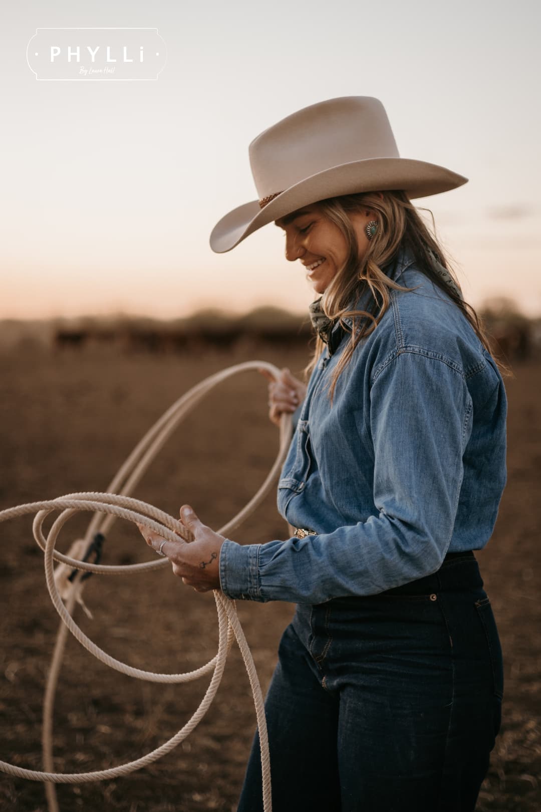 Model wearing the beige cowboy hat Wheeler Beige by PHYLLi Designs with chocolate leather hat band while handling a lariat in a dusty paddock at sunset.