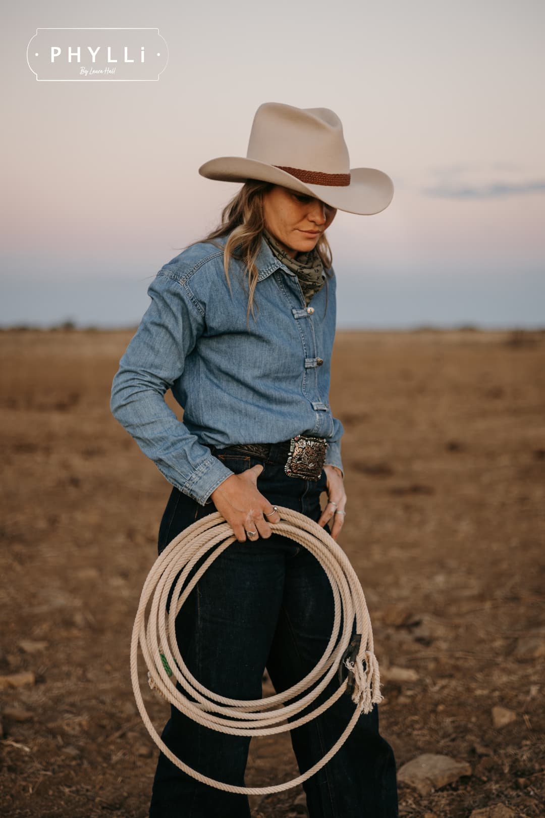 Model wearing the beige cowboy hat Wheeler Beige by PHYLLi Designs with chocolate leather hat band holding a stockyard lariat.