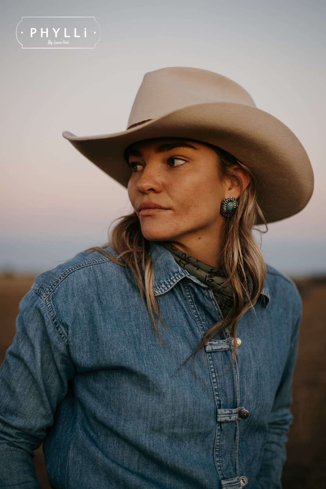 Model wearing the beige cowboy hat Wheeler Beige by PHYLLi Designs with chocolate leather hat band looking across an open paddock.
