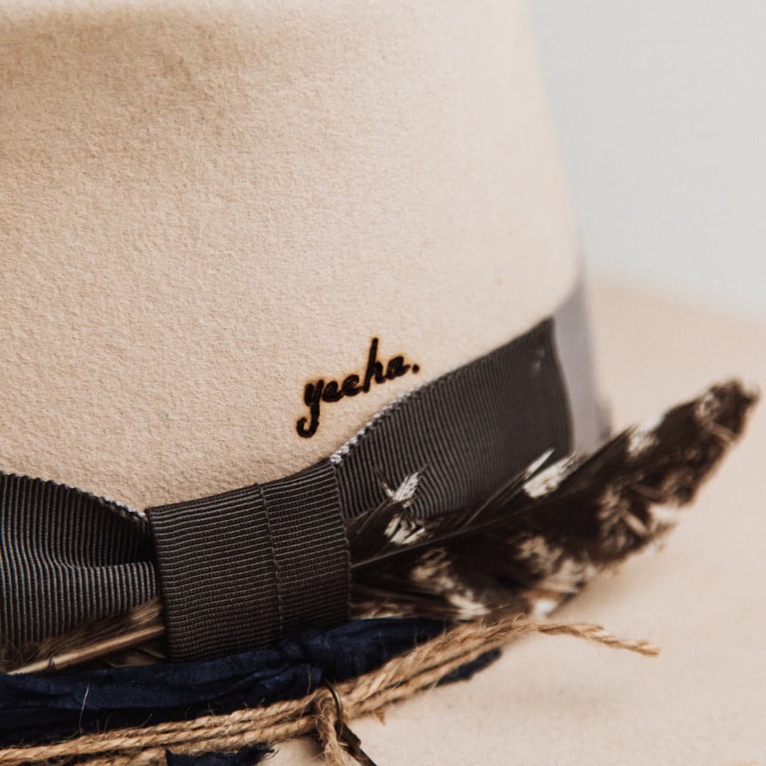 Close up detail of the Wrangler beige distressed felt hat by PHYLLi Designs showing the steel grey grosgrain bow with layered feathers and royal blue sari silk trim.