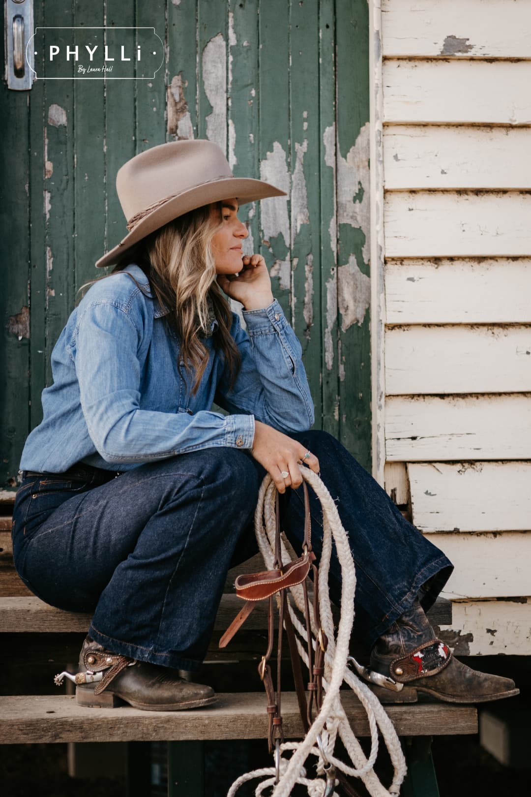 Woman wearing the Sahara cowboy hat Wrapped Wheeler Sahara by PHYLLi Designs with tonal tie-dye sari silk wrap resting on shed steps in a western denim outfit.