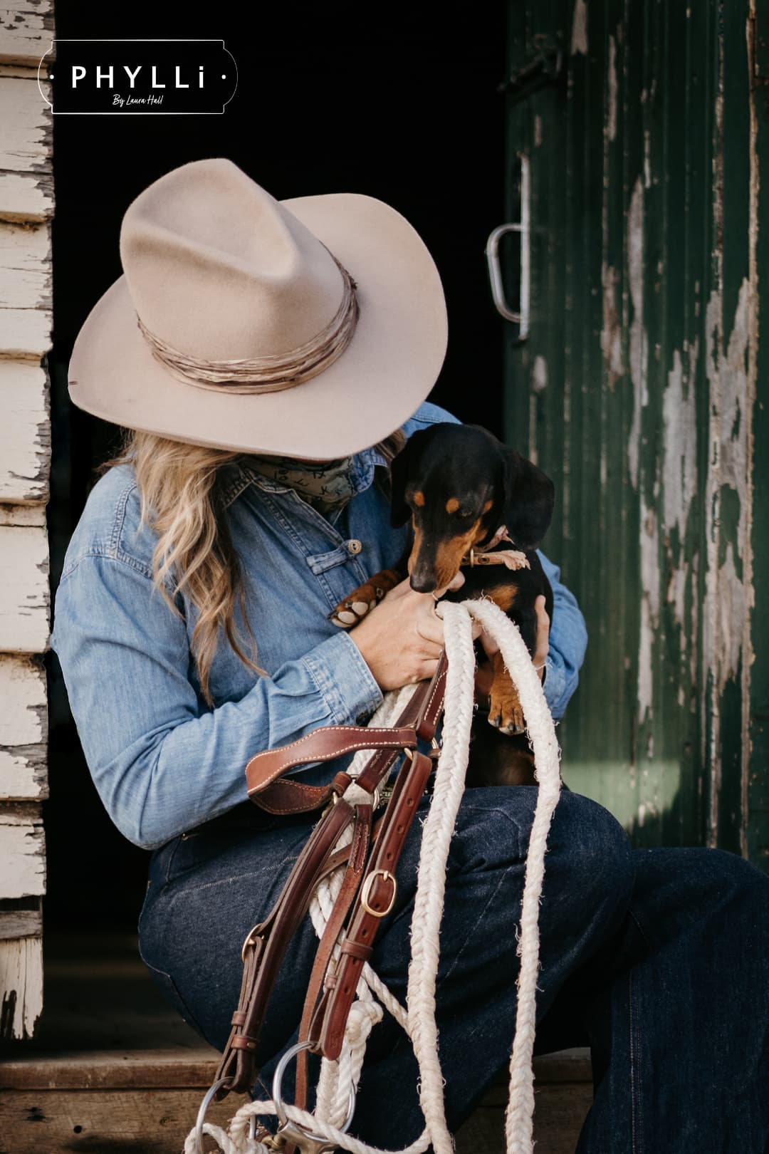 Woman wearing the Sahara cowboy hat Wrapped Wheeler Sahara by PHYLLi Designs with tonal tie-dye sari silk wrap sitting on wooden steps holding a dachshund and stockman rope outside a rustic shearing shed.