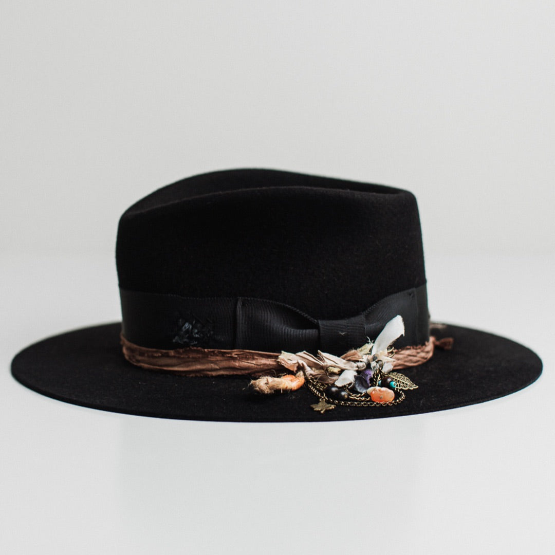 Left side view of the Newmarket dark navy felt hat by PHYLLi Designs with bronze chunky hat pin featuring sari silks, tassel, charms and beads.