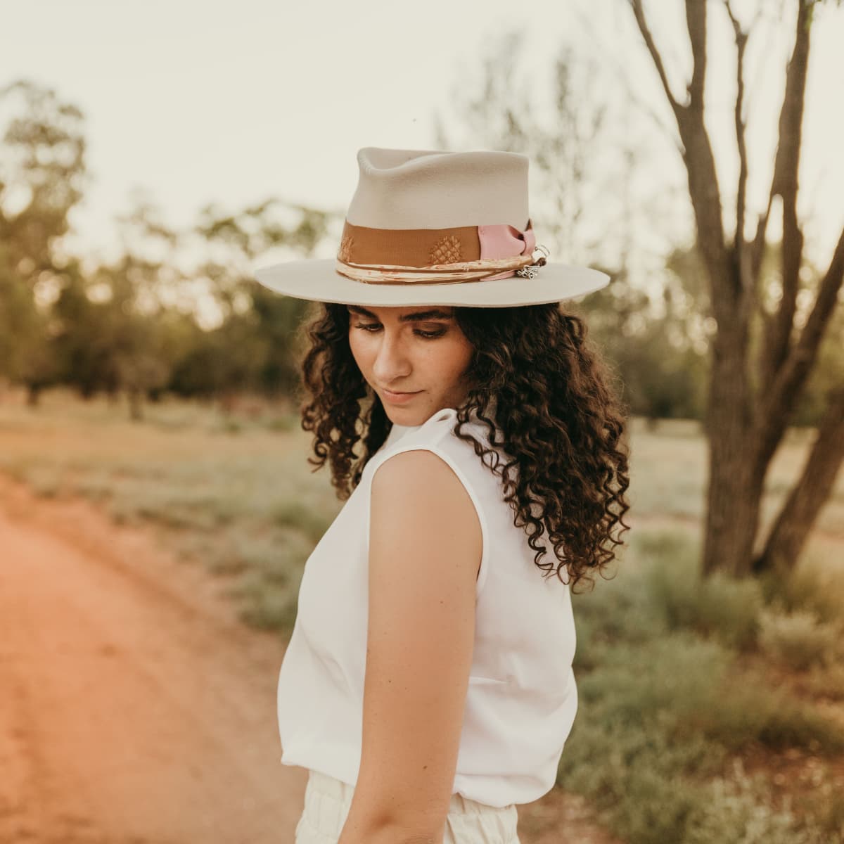 Sarah Wheeler wearing a beige hat with brown band and decorative elements, standing on a dirt path in a natural setting.