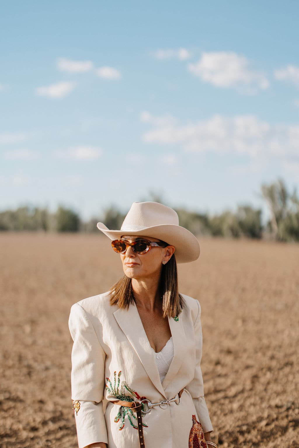 Woman wearing the PHYLLi B&W Wheeler beige cowboy hat and a linen jacket, standing in a paddock under a blue sky.