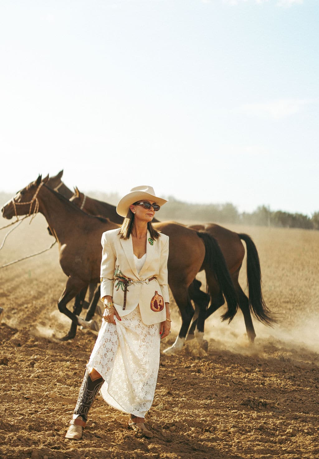 Woman wearing the PHYLLi B&W Wheeler beige cowboy hat and a white outfit, standing in a paddock with horses at sunset.