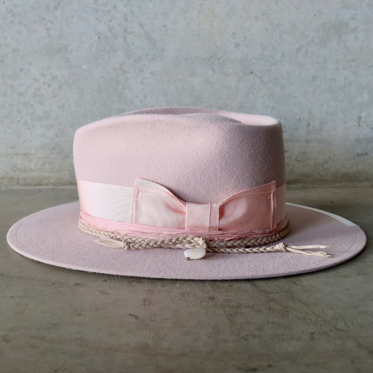 Left side view of the baby pink felt hat Argyle Limited Edition by PHYLLi Designs featuring plaited nude suede string with rough pearl pendant.