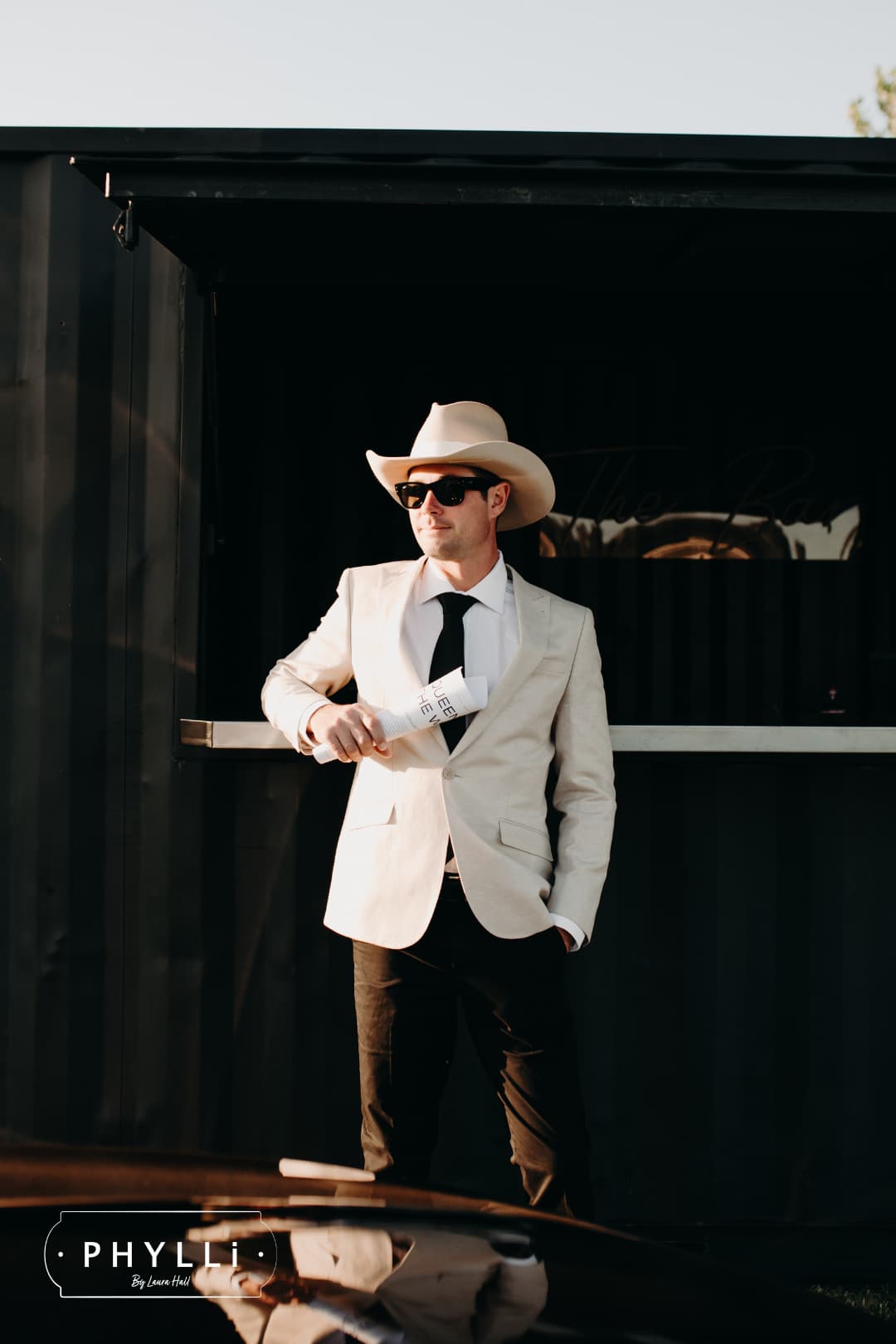Male model wearing the beige felt cowboy hat B&W Wheeler by PHYLLi Designs with ivory Japanese grosgrain band at an outdoor bar setting.