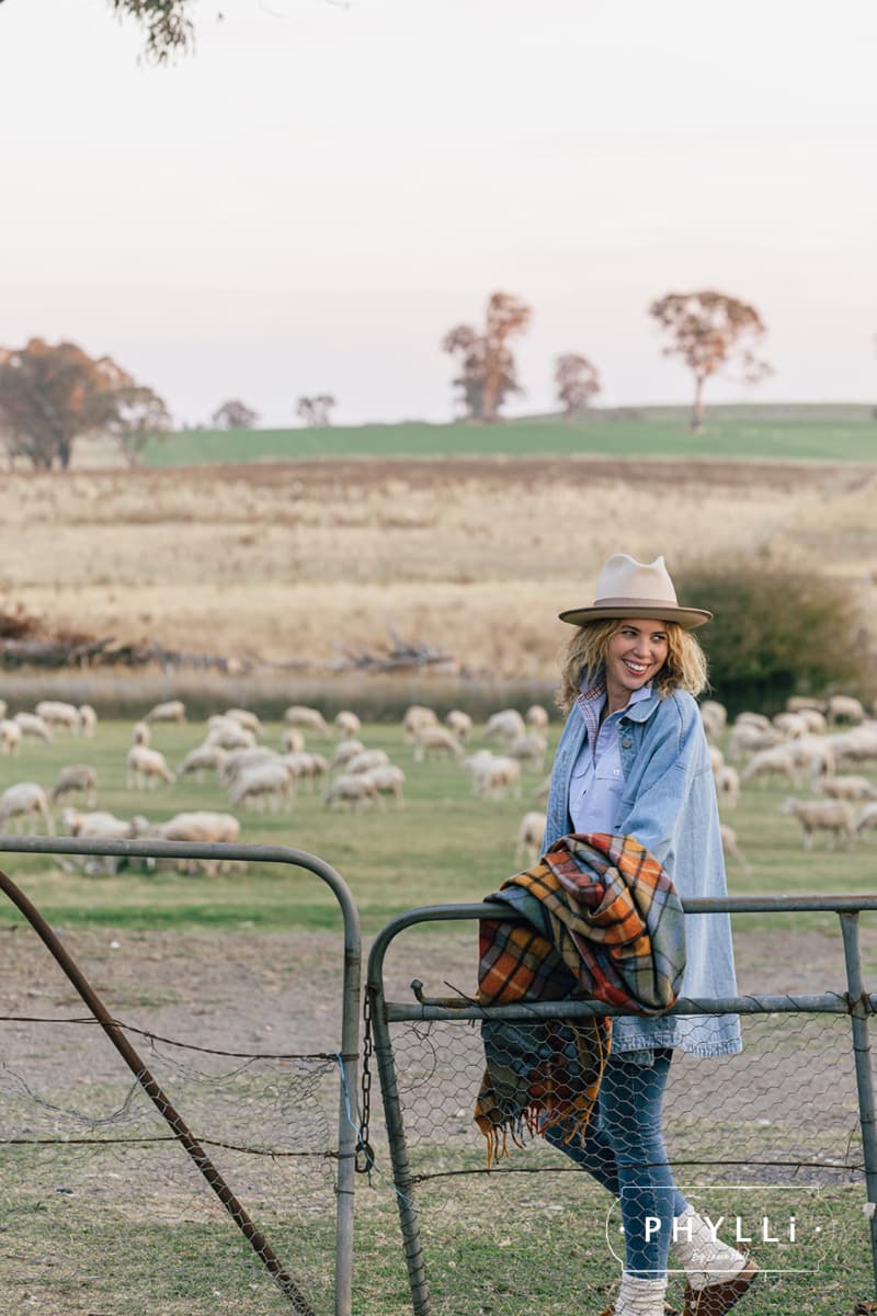 Model wearing the Buckler beige felt hat by PHYLLi Designs with taupe grosgrain trim, denim trimming and twine plait in a rural paddock with sheep.