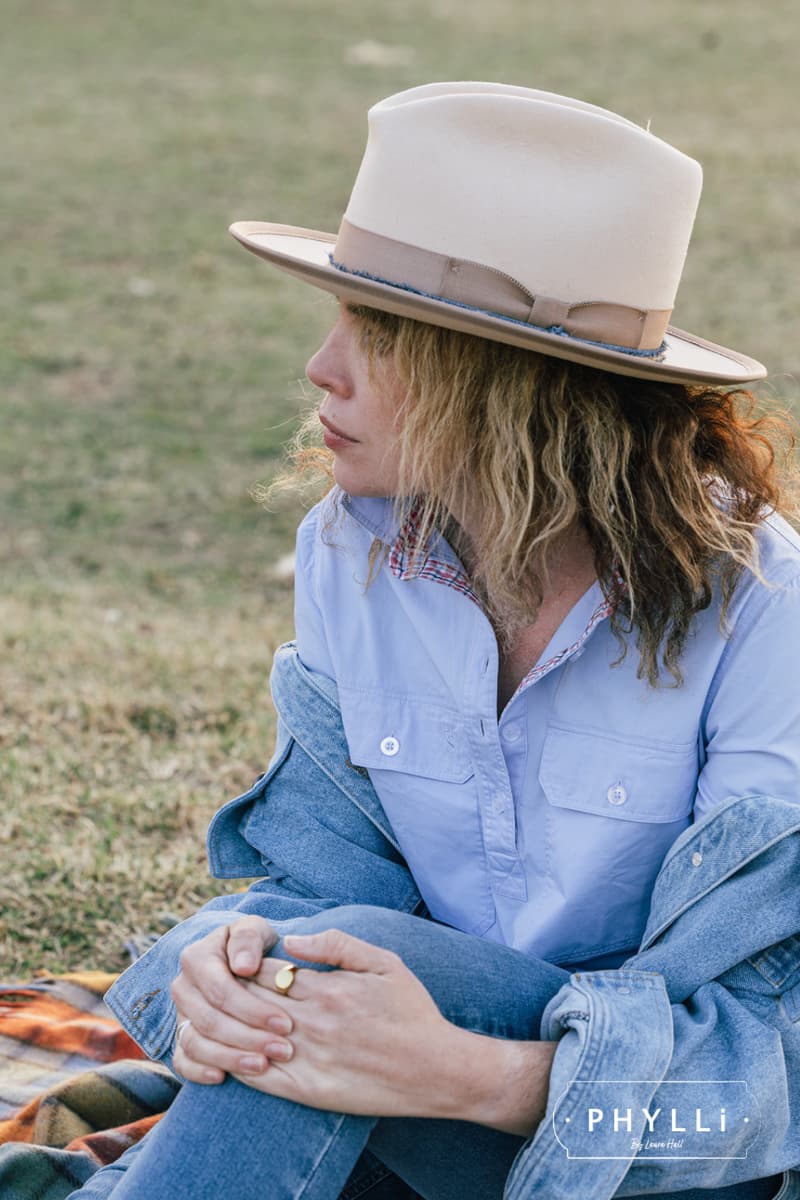 Model wearing the Buckler beige felt hat by PHYLLi Designs with taupe grosgrain trim, denim trimming and twine plait while seated outdoors on grass.