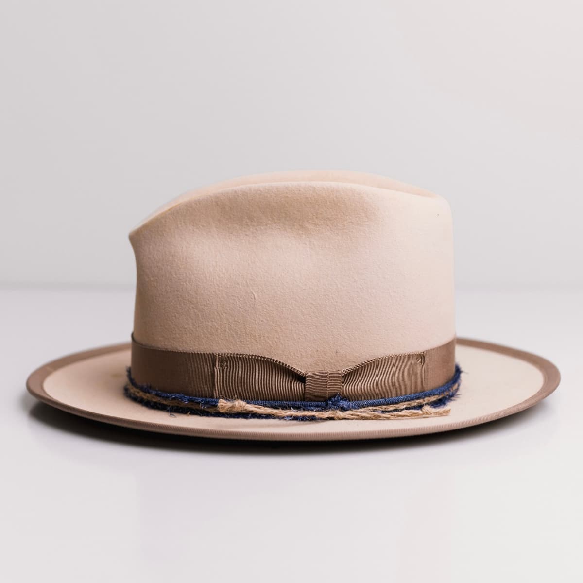 Left side view of the Buckler beige felt hat by PHYLLi Designs with taupe Japanese grosgrain trim, denim trimming and twine plait.